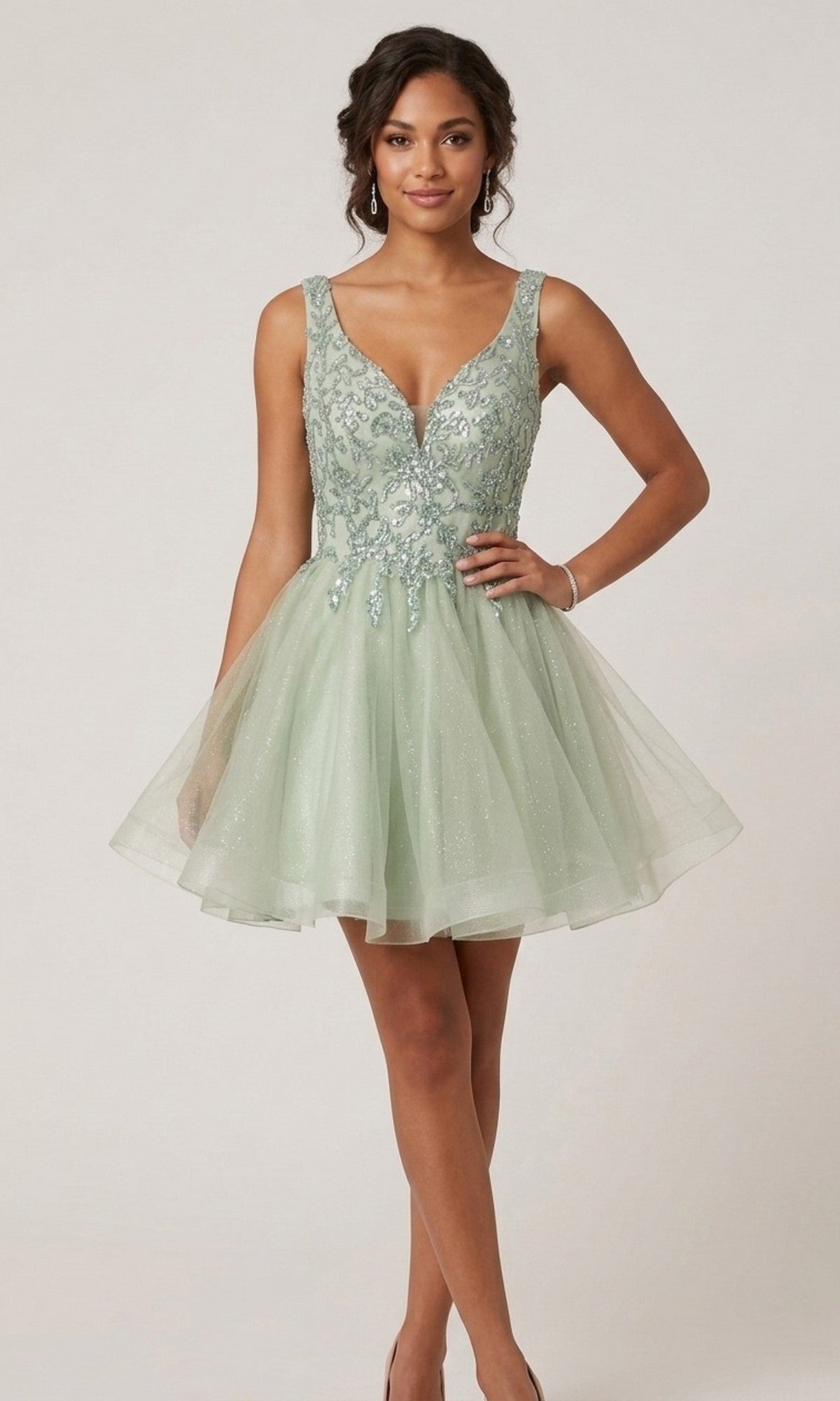 A woman wearing the Chicas C376 Short Homecoming Dress in sage—a sleeveless, V-neck fit-and-flare style crafted from glitter tulle with delicate floral embroidery—stands against a plain background.